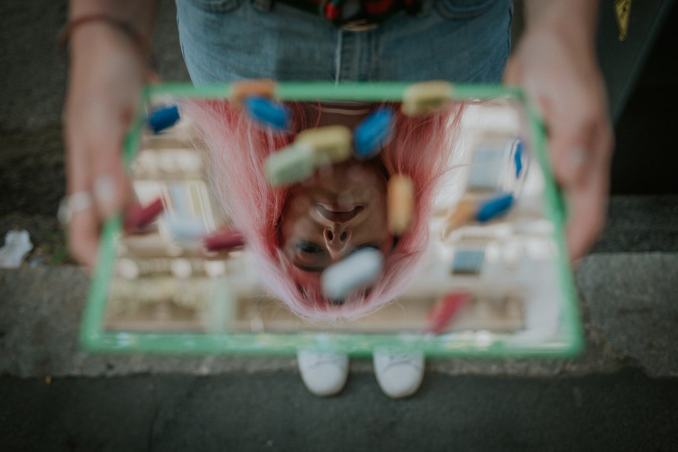 Pink haired woman looking down into a hand-held mirror with various pill capsules on it.